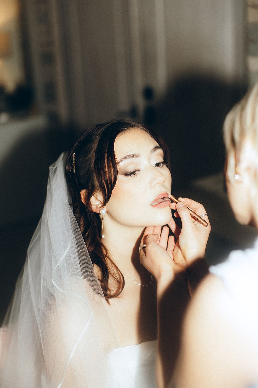 Professional makeup artist applies lipstick to a bride wearing a white veil before her wedding ceremony.