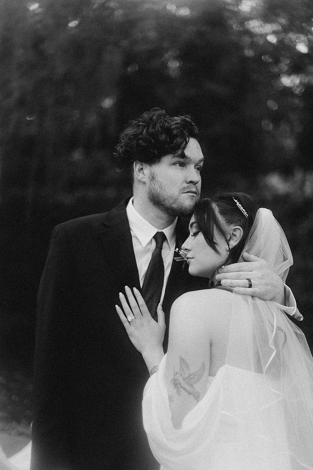 Black and white portrait of bride and groom embracing, bride in veil resting head on groom's shoulder.
