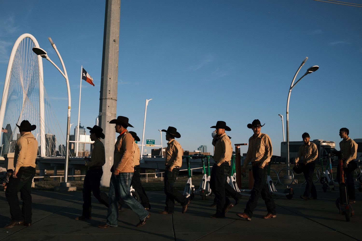 Cowboys in tan shirts and hats march across a bridge with a Texas flag and city skyline in the background.