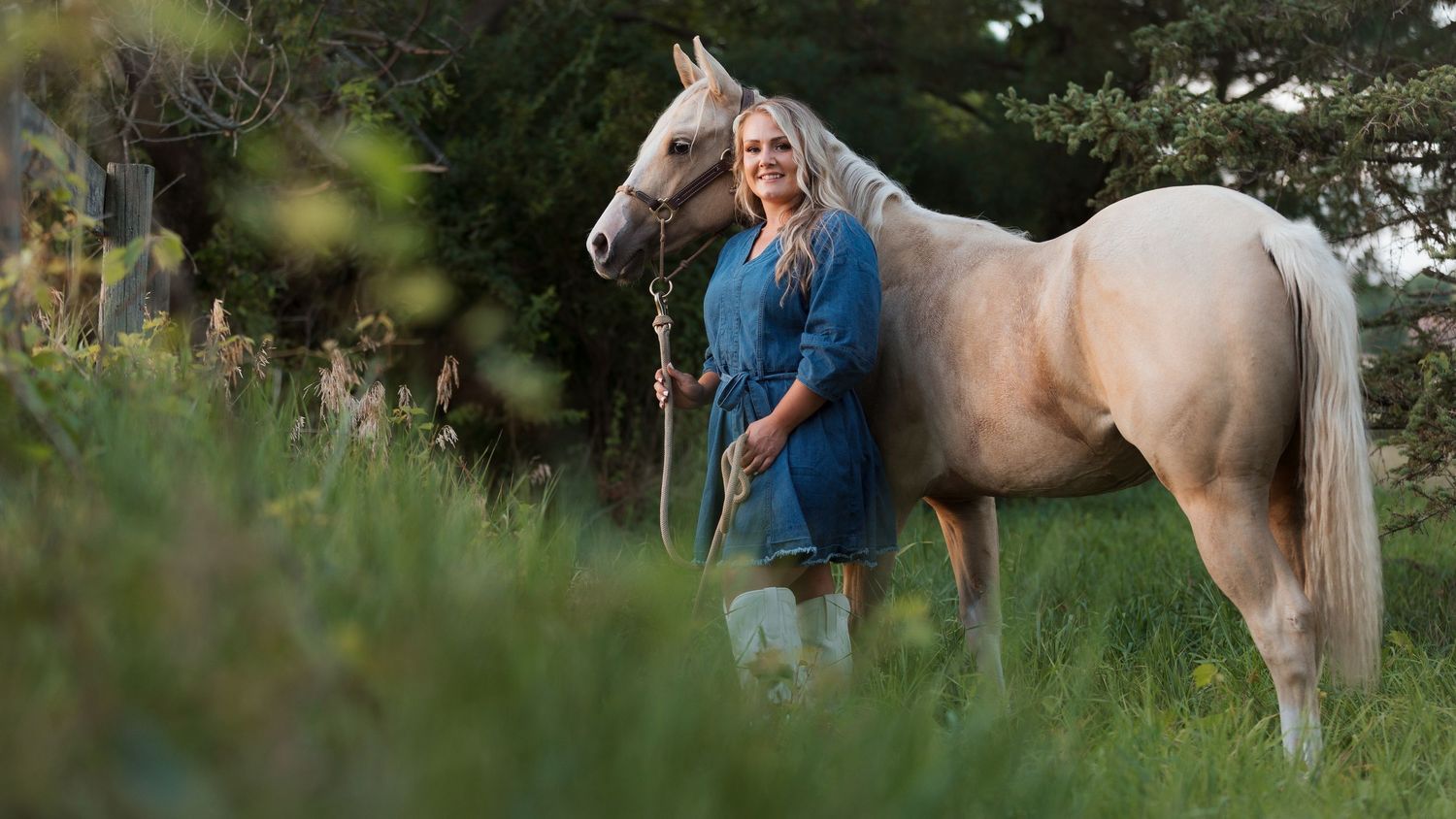 Woman in blue dress standing beside a palomino horse in a lush green meadow surrounded by trees.