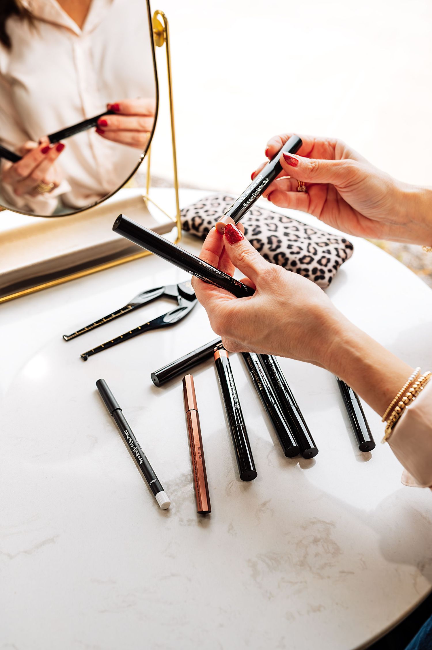 Close-up of hands holding makeup pencils on marble vanity table with beauty tools and mirror.