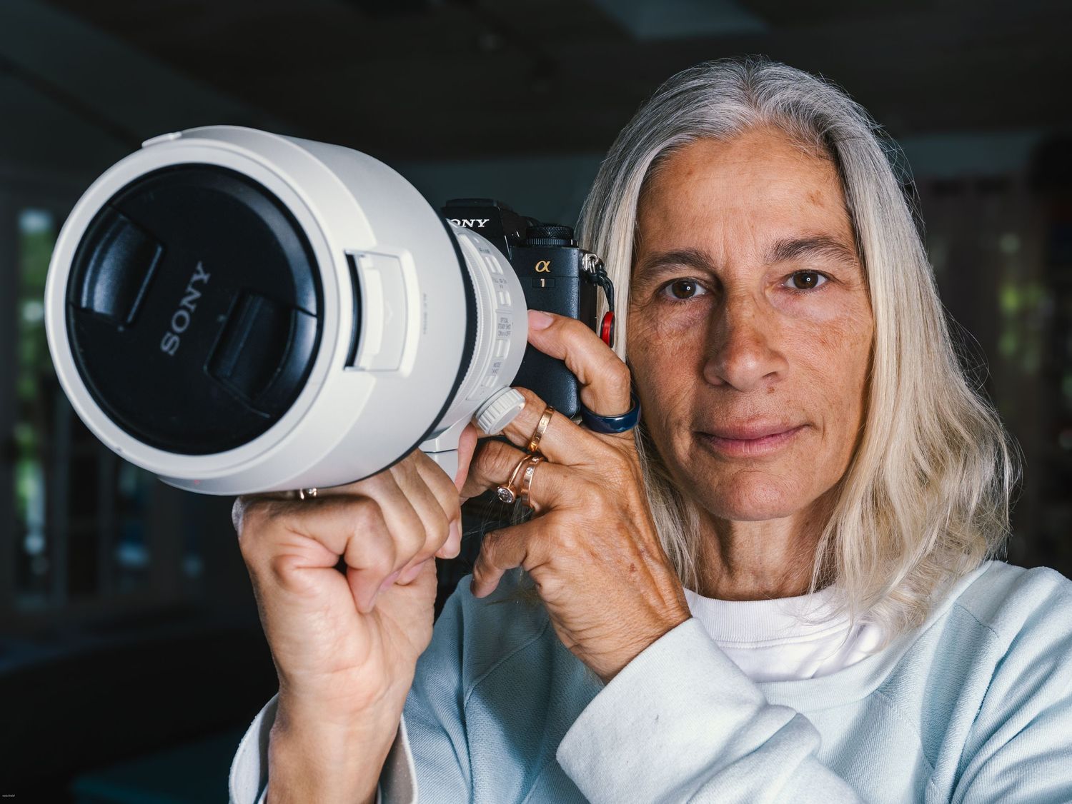 A woman with gray hair holds a Sony camera with a white telephoto lens, posing confidently indoors.
