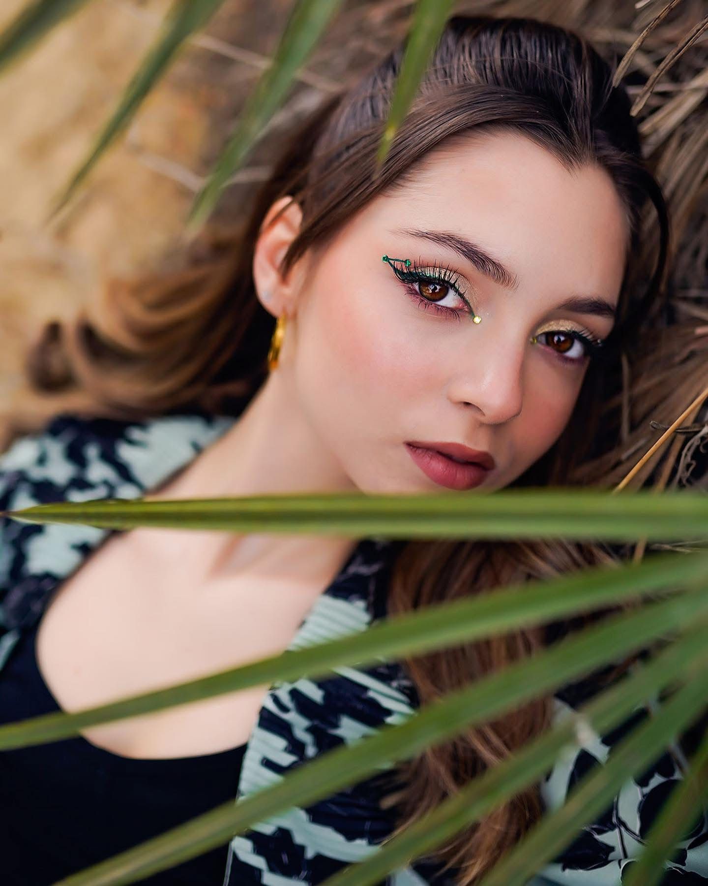 Belle jeune femme avec un maquillage coloré des yeux allongée parmi les feuilles de palmier tropical sous la chaude lumière du soleil.