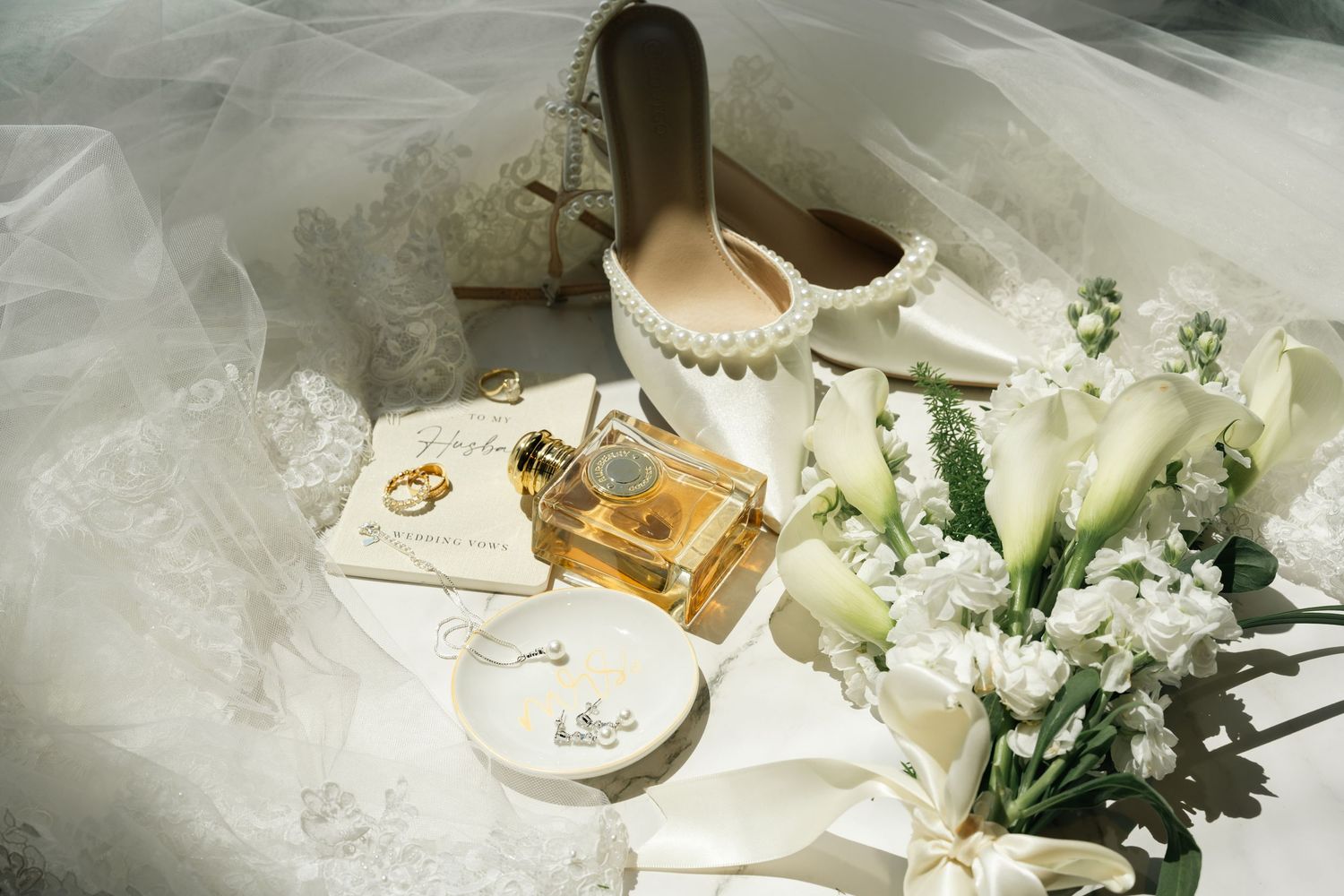 Elegant bridal flat lay featuring pearl heels, gold perfume bottle, wedding rings, and lace veil on white background.
