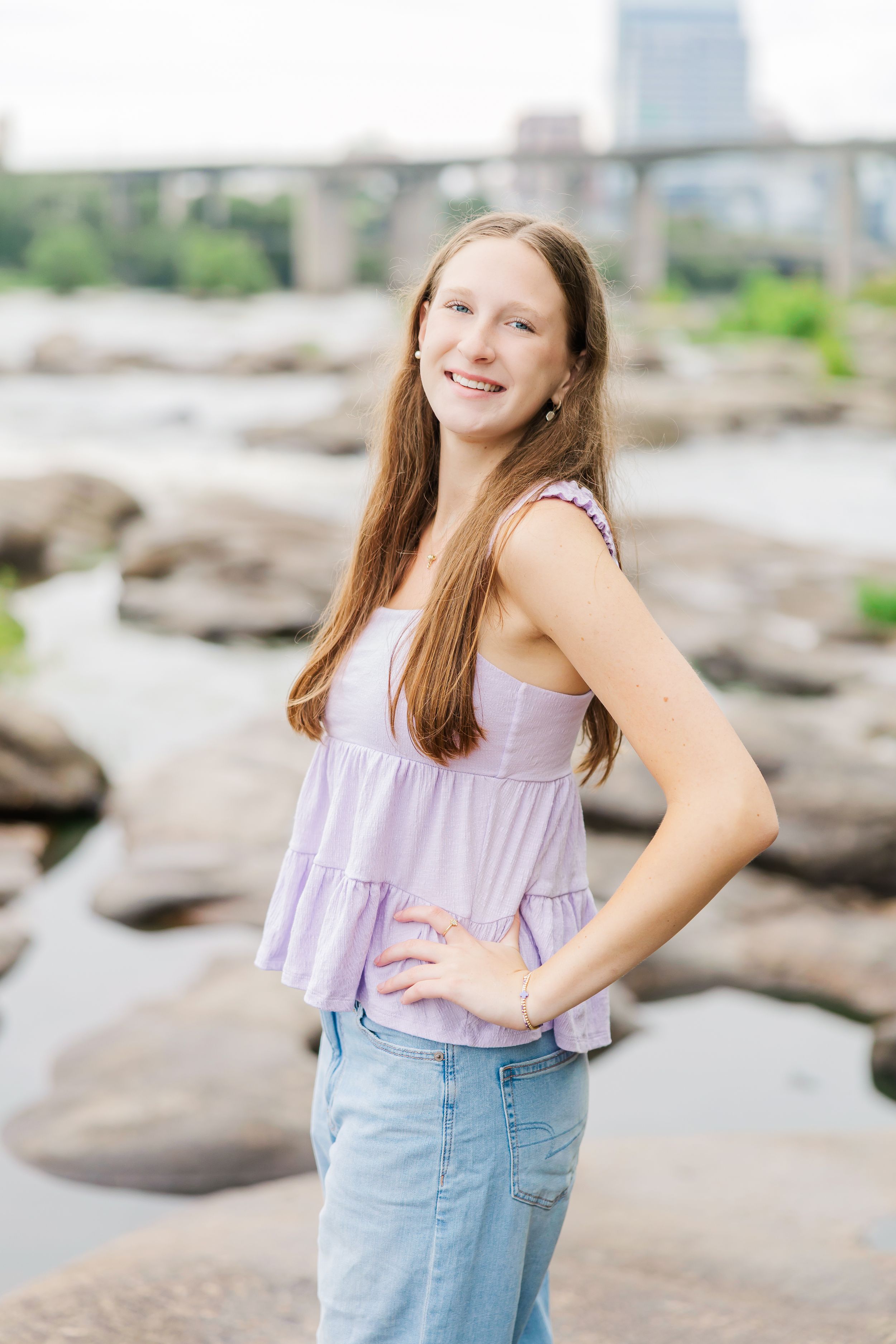 Smiling teenage girl in lavender top and jeans poses on rocky riverbank with bridge in background.