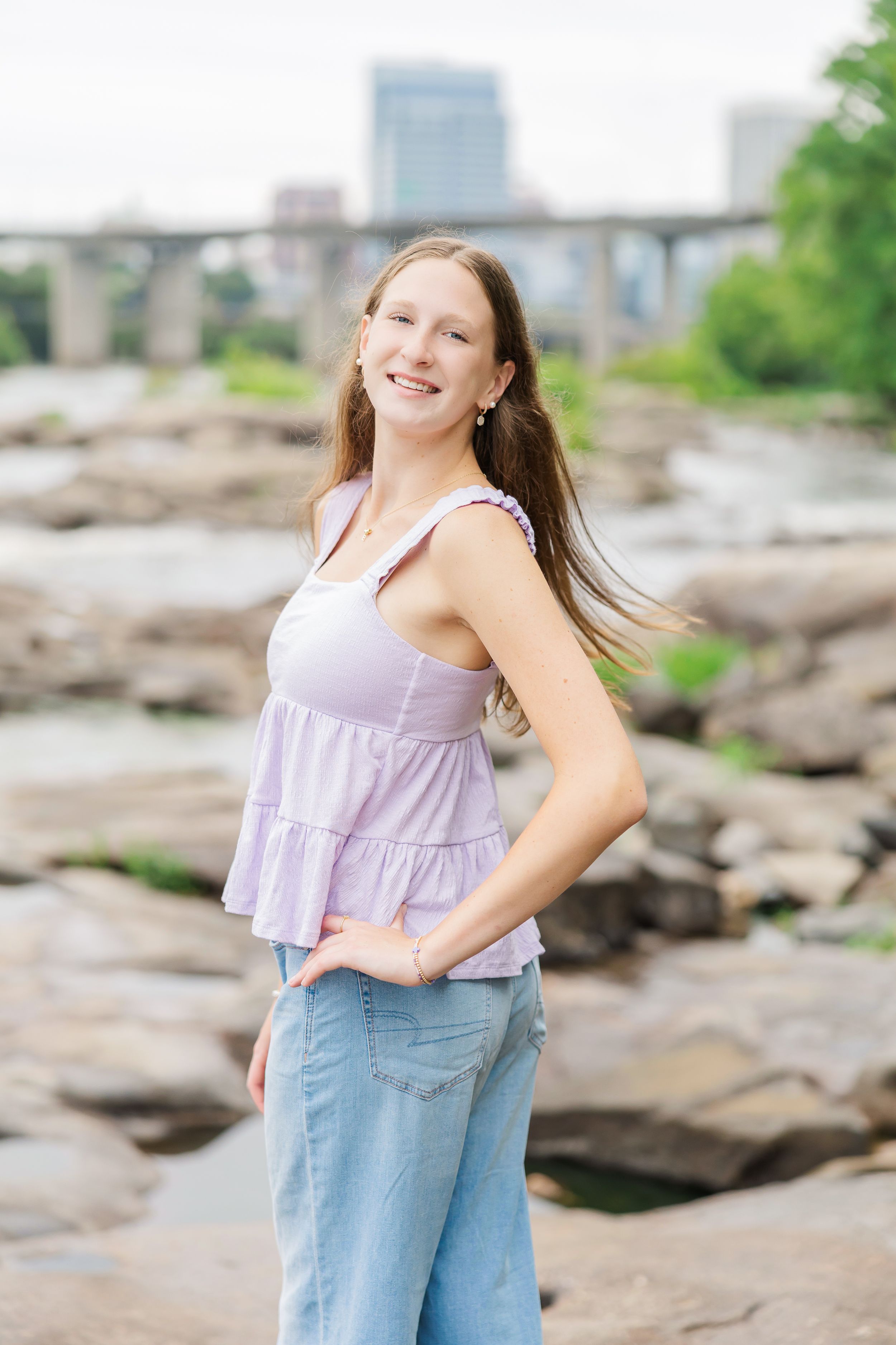 Smiling teen girl in lavender top and wide-leg jeans poses on rocks by a river with a bridge backdrop.