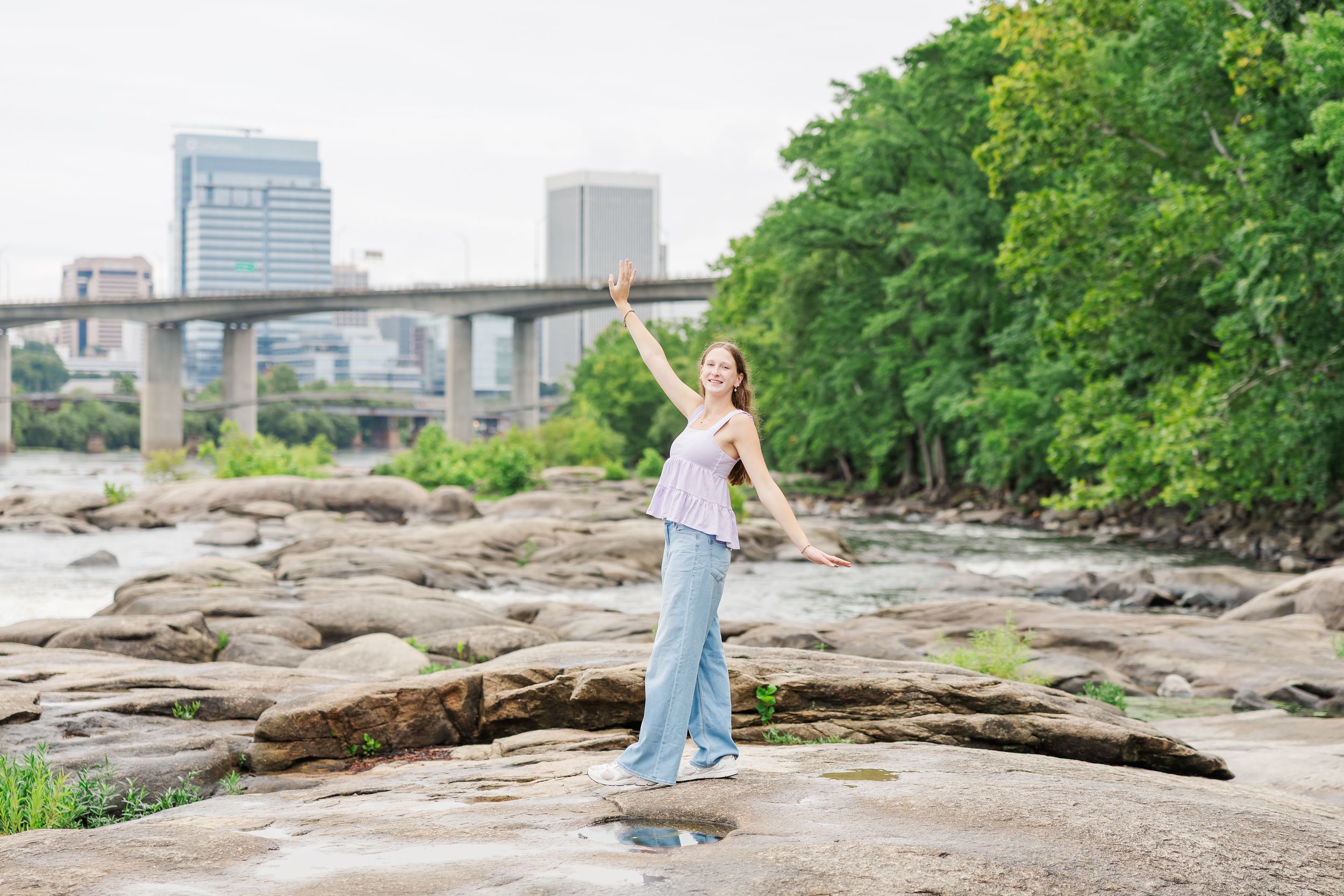 A joyful woman stands on rocky riverbank with one arm raised, city skyline and bridge visible in background.