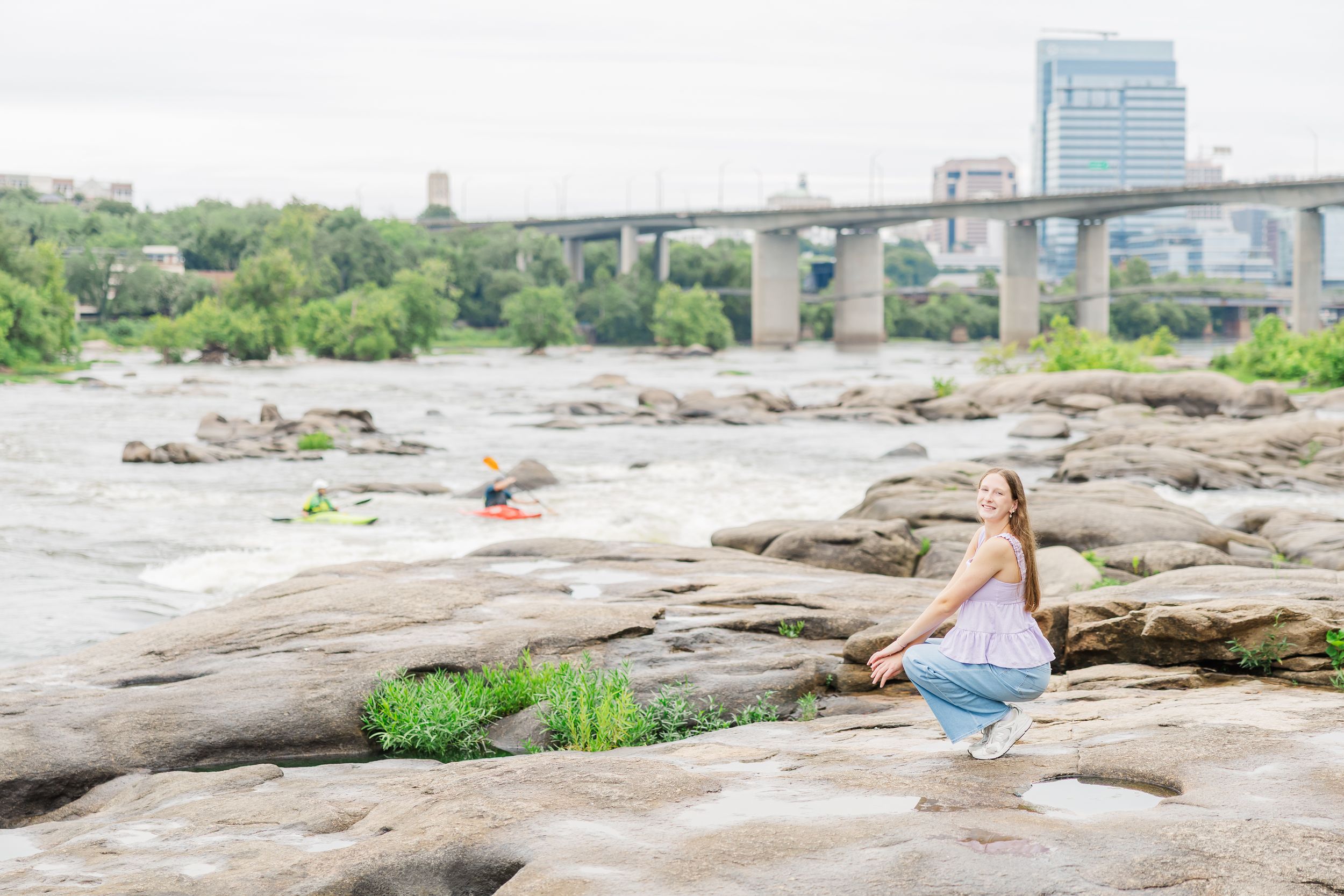 A young girl crouches on rocky riverbank with a bridge and city skyline visible in the background.