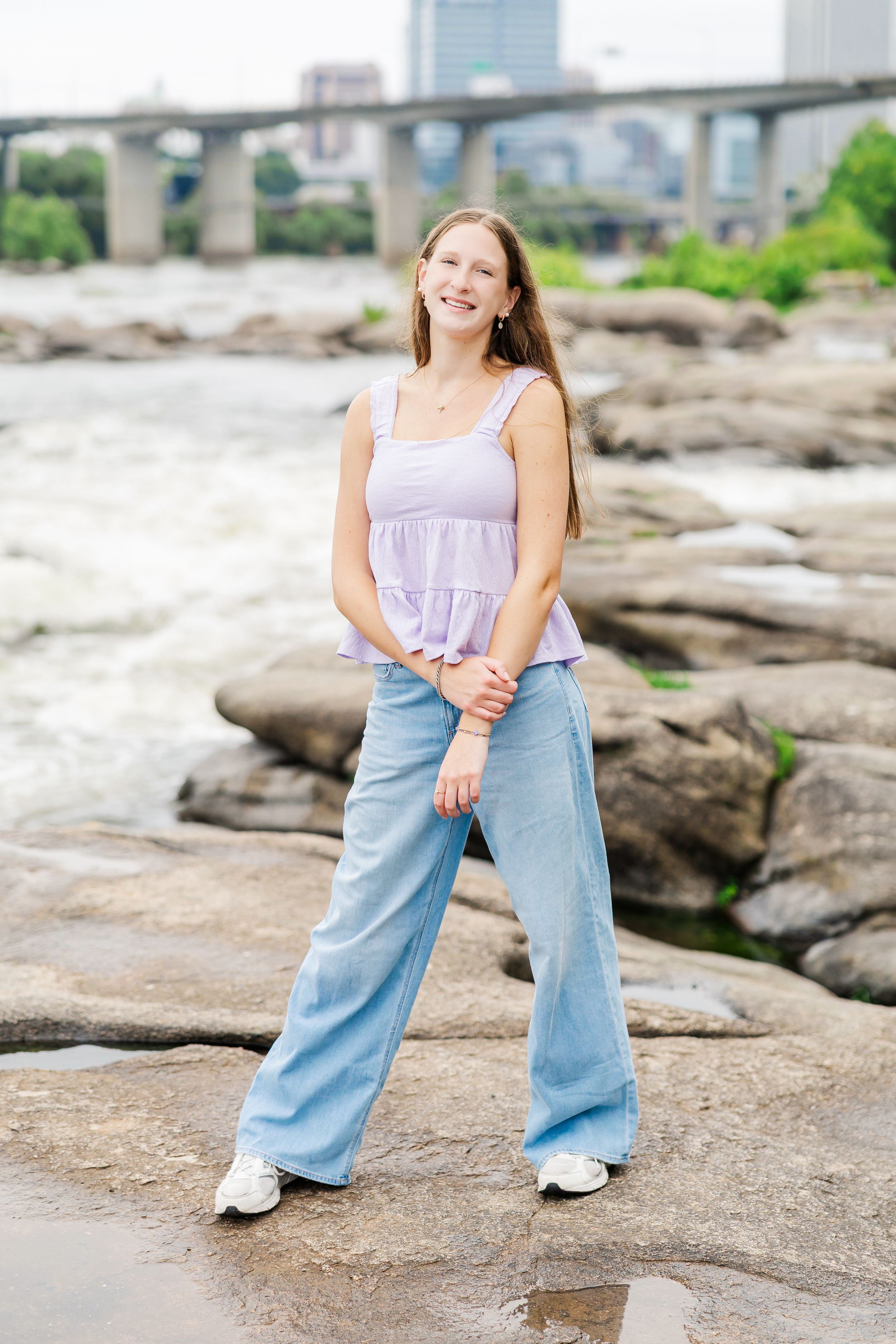 Young woman in purple tank top and wide-leg jeans stands on riverside rocks smiling with city bridge behind her.