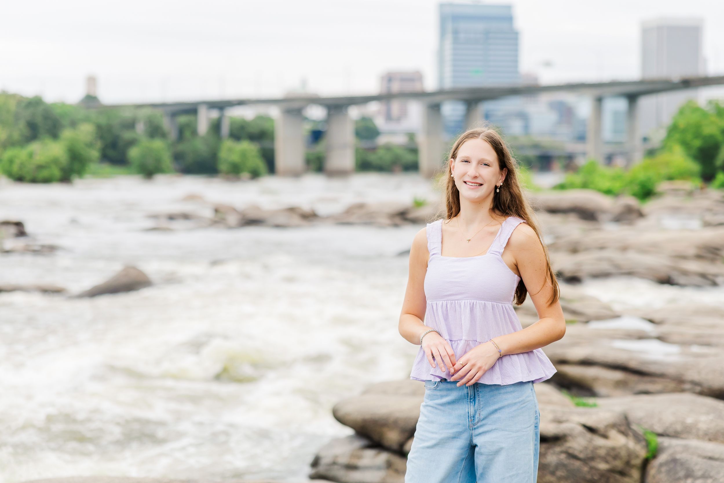 Young woman smiling by rocky rapids with a bridge and city skyline in the background.