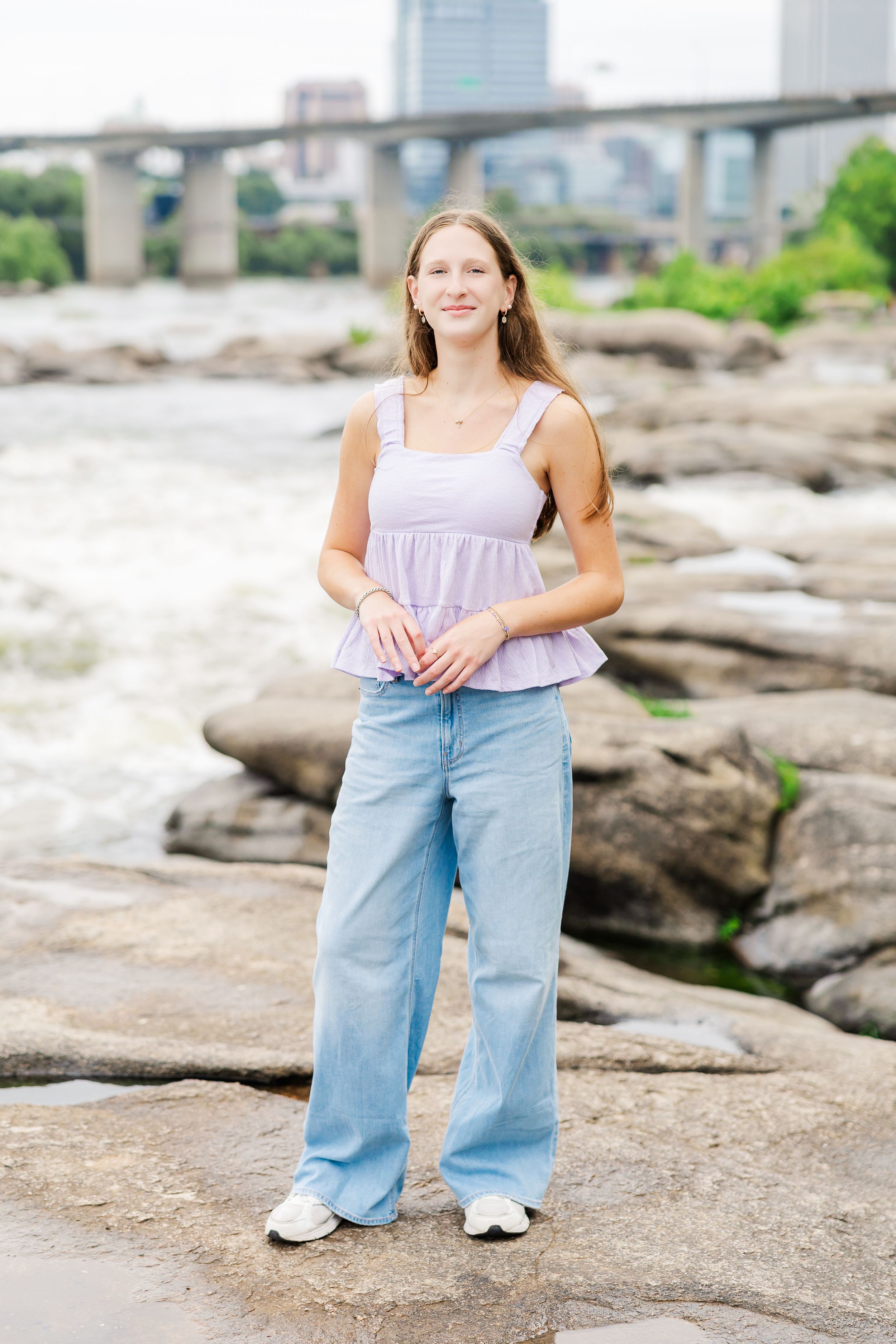 Young woman in lavender top and wide-leg jeans standing on rocks by a river with a city bridge backdrop.