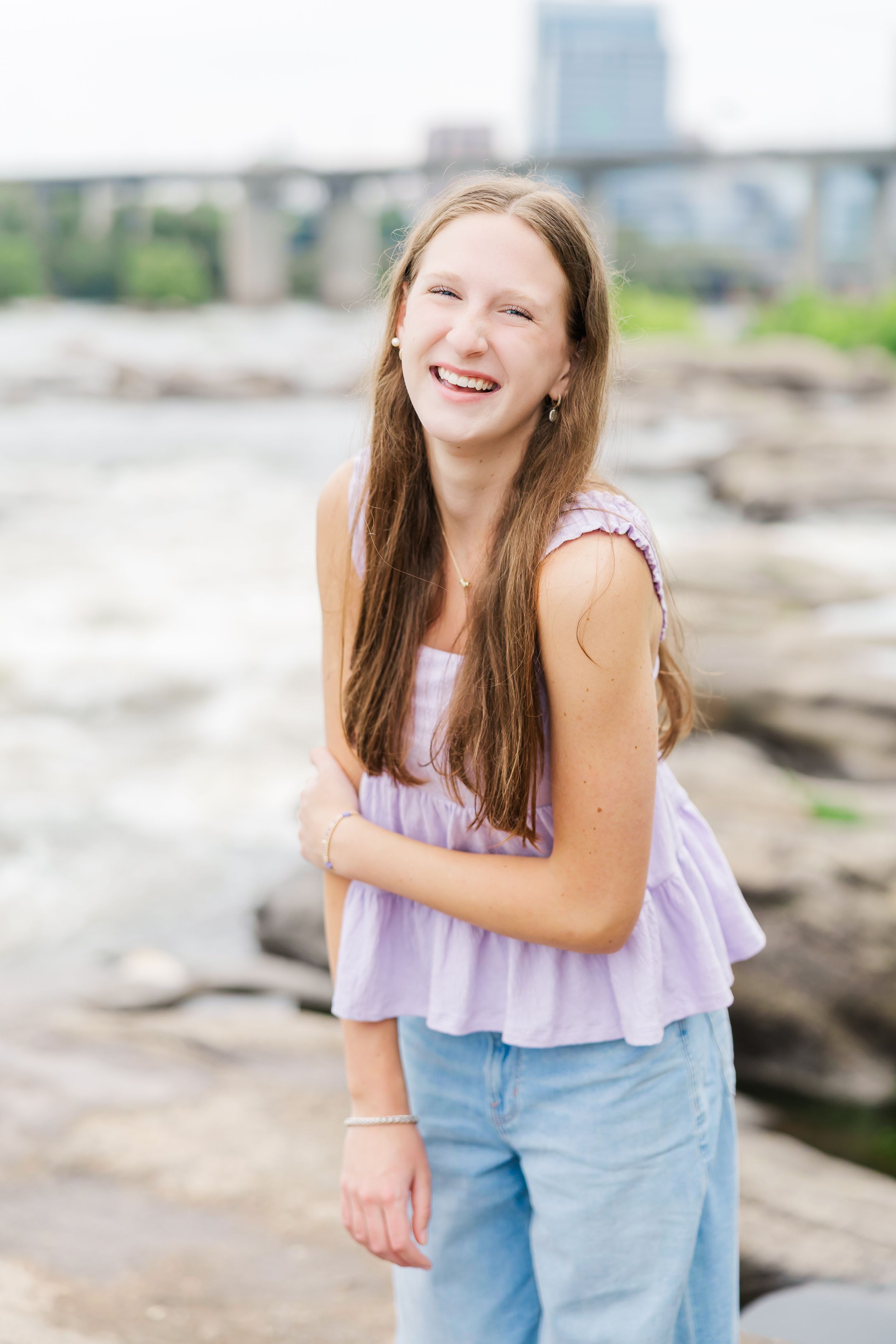 Smiling teenage girl with long brown hair wearing a lavender top and jeans, posing by a rocky riverbank.