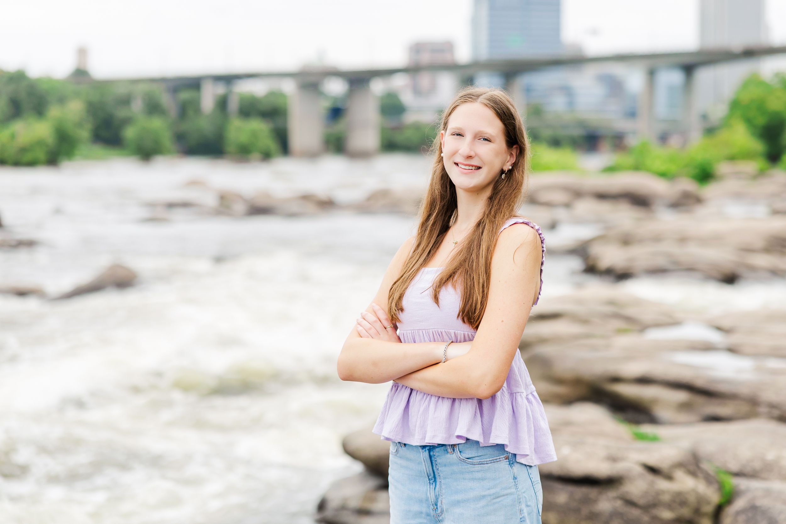 Teen girl smiling with arms crossed on rocky riverbank, bridge and city skyline in background.