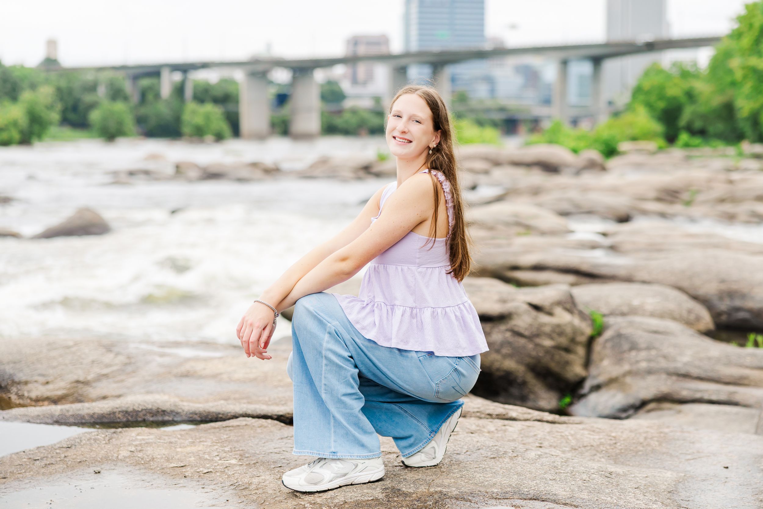 Young woman in pink top and jeans crouching on rocks by a river with a bridge in the background.