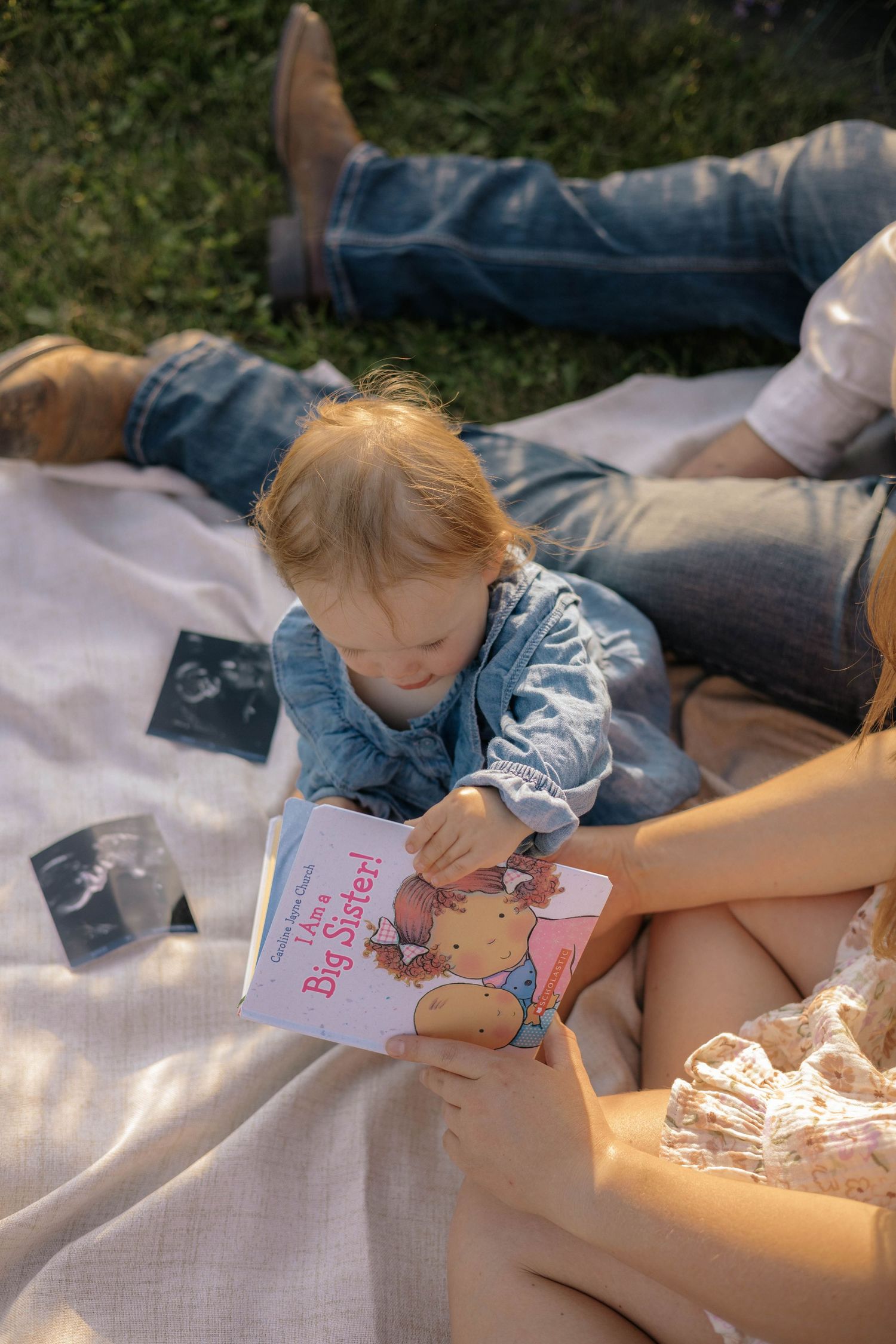 Toddler reading I'm a Big Sister book on blanket with parents and ultrasound photos.