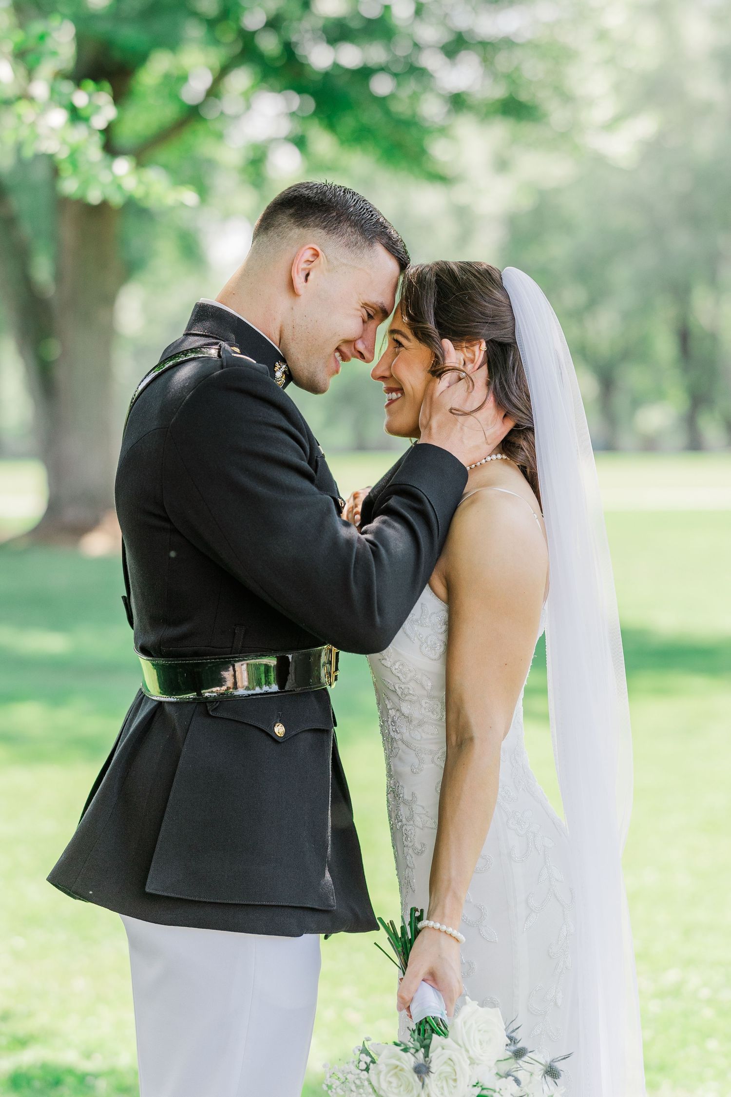 Military groom in dress uniform tenderly holds bride's face as they share an intimate moment outdoors.