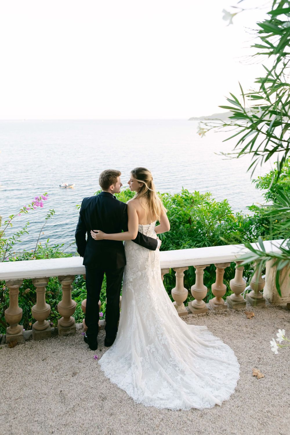 Bride and groom embracing on elegant balcony overlooking serene blue sea, surrounded by lush greenery on their wedding day.