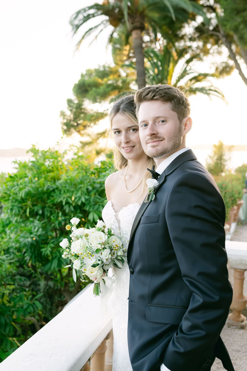 Bride and groom pose on a sunlit balcony with lush greenery, bride holding a white bouquet.