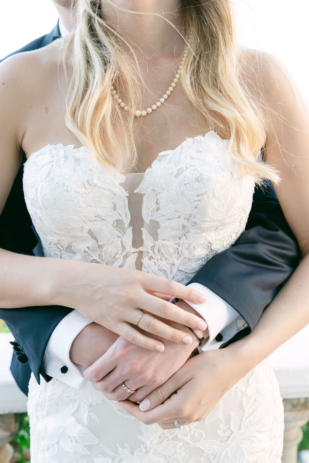 Bride in lace strapless gown with pearl necklace, groom embracing her from behind, hands clasped showing wedding rings.
