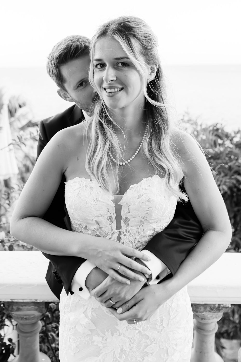 Black and white photo of a smiling bride in lace gown and pearl necklace embraced by groom in suit outdoors.
