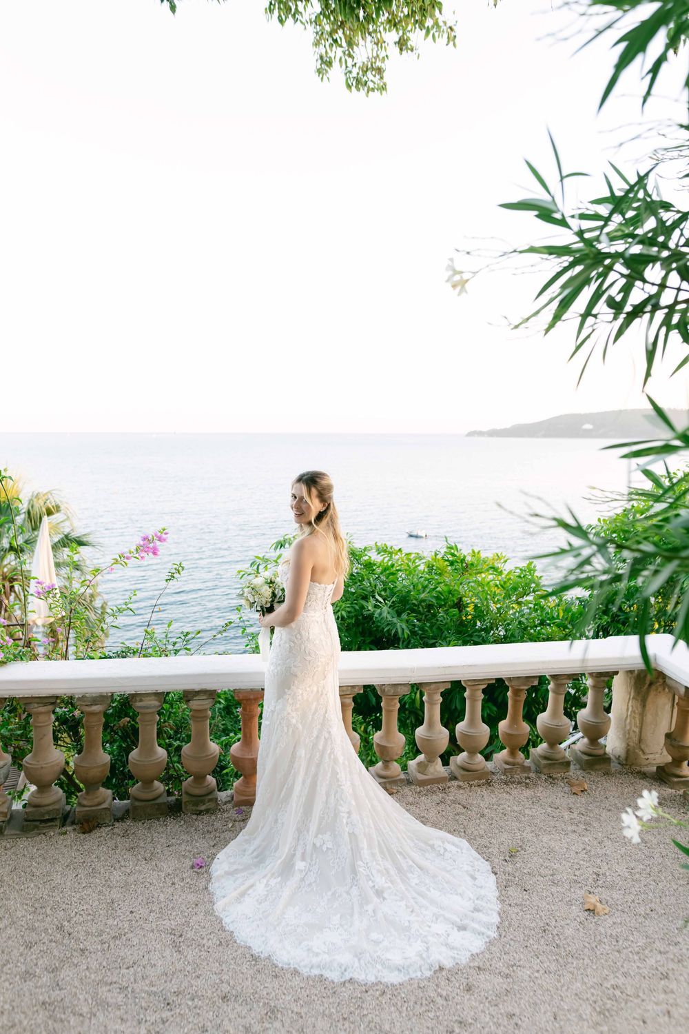 Bride in lace wedding gown with train stands on elegant terrace overlooking serene blue sea surrounded by lush greenery.