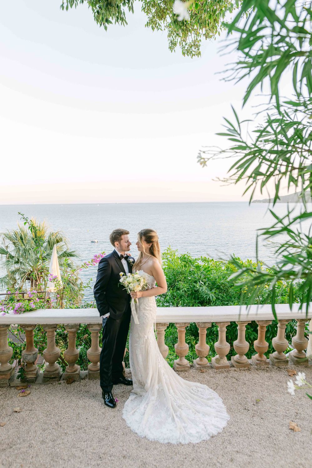 Bride and groom embrace on elegant stone balcony overlooking the sea, surrounded by lush greenery at sunset.