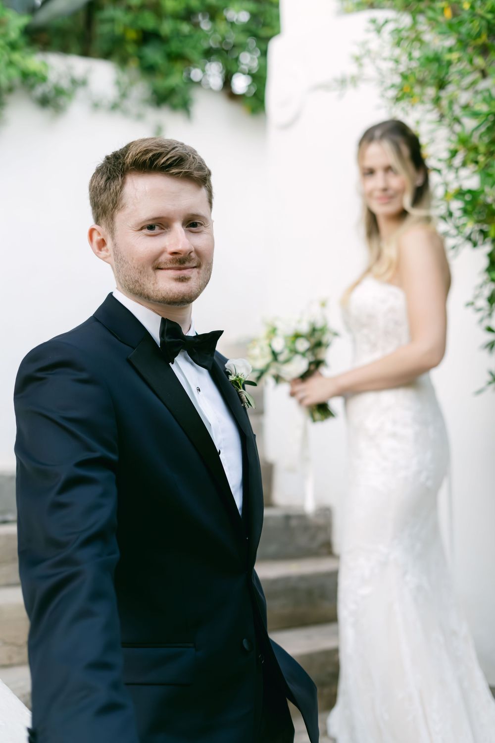 Groom in black tuxedo smiling at camera with bride in white lace gown holding bouquet standing behind him.