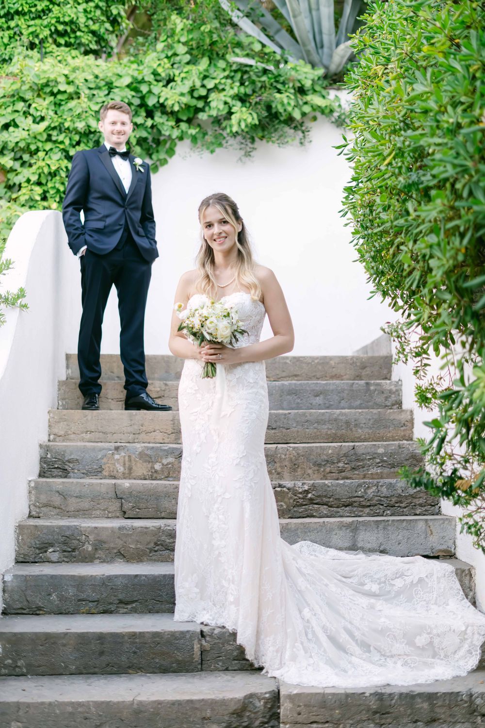 Bride in lace mermaid gown holds bouquet on stone steps while groom in black tuxedo stands behind her.