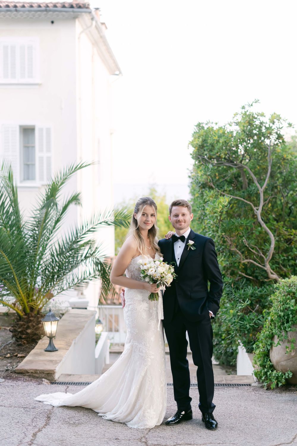Wedding couple poses together outside white villa, bride in strapless lace gown holding white bouquet.