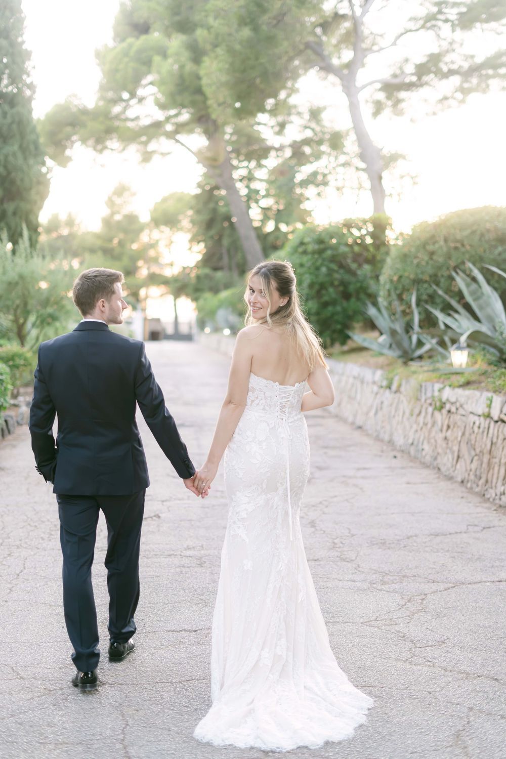 Bride and groom walk hand in hand down a sunlit garden path, she in a strapless lace gown, he in a navy suit.