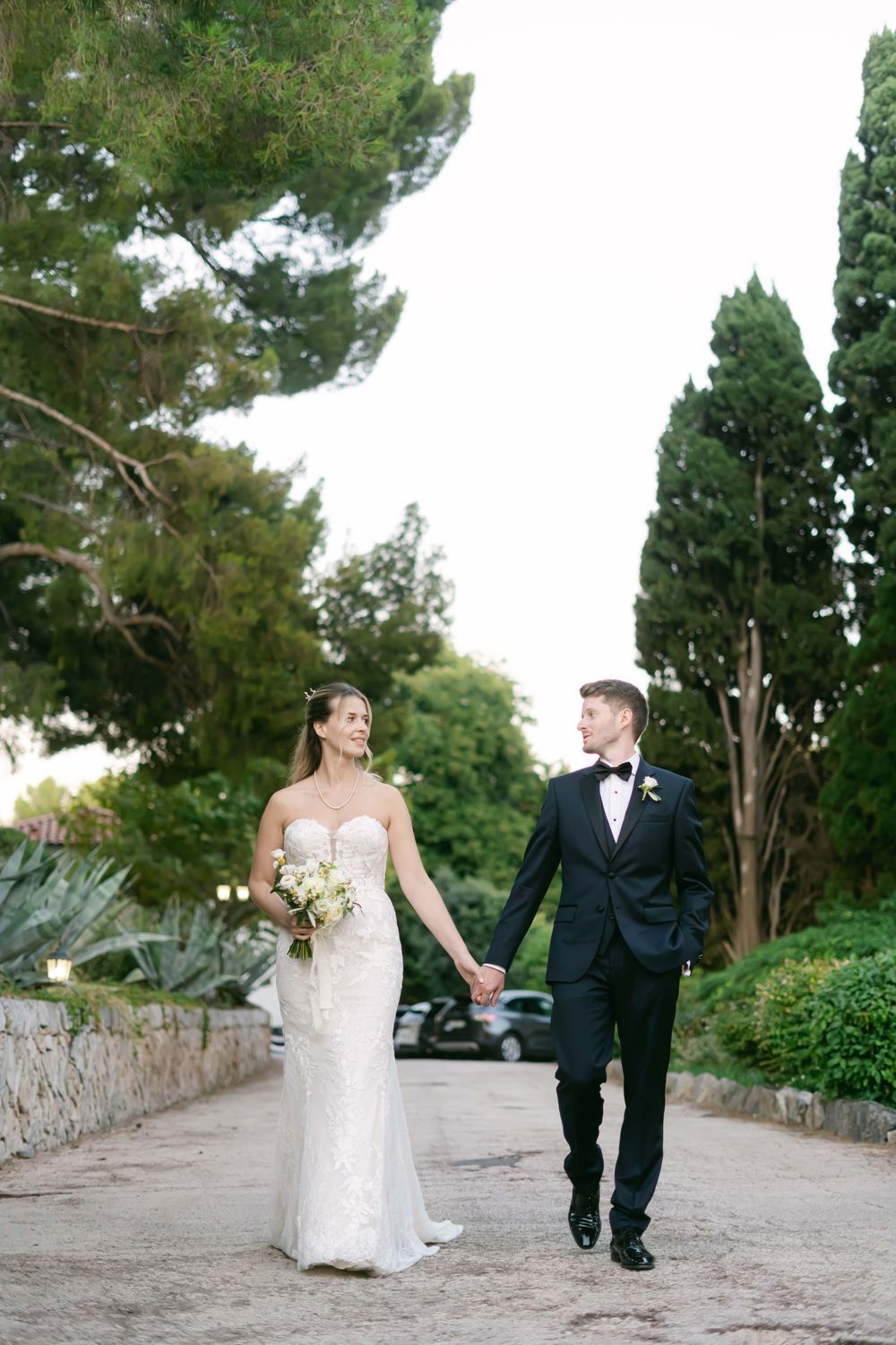 Bride in white gown and groom in black tuxedo hold hands walking along a garden path lined with tall trees.