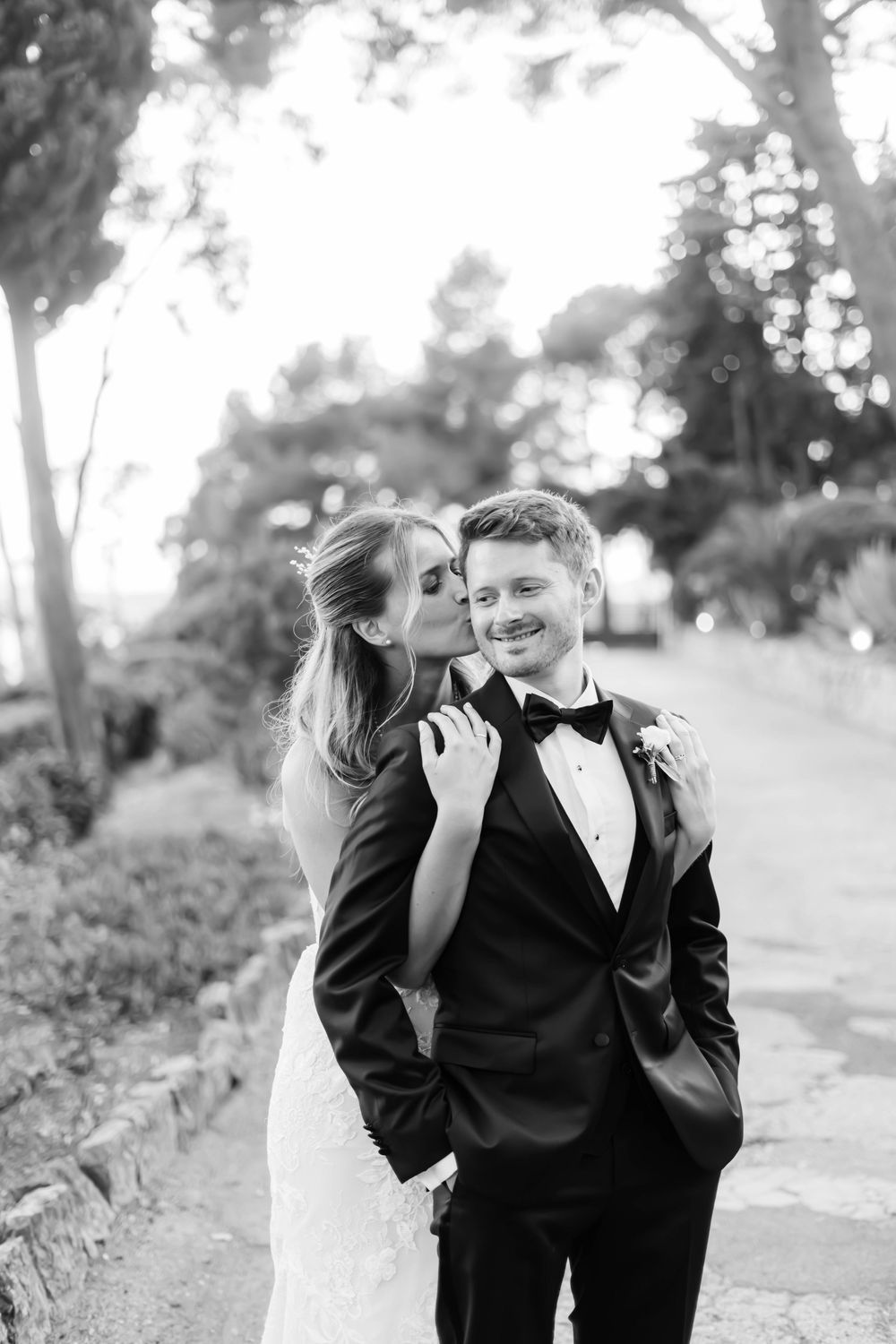 Black and white wedding photo of bride kissing groom on cheek outdoors, groom smiling in tuxedo with boutonniere.