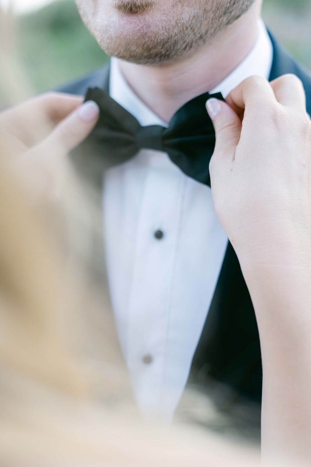 Hands adjusting a black bow tie on a groom wearing a classic black tuxedo with white dress shirt.