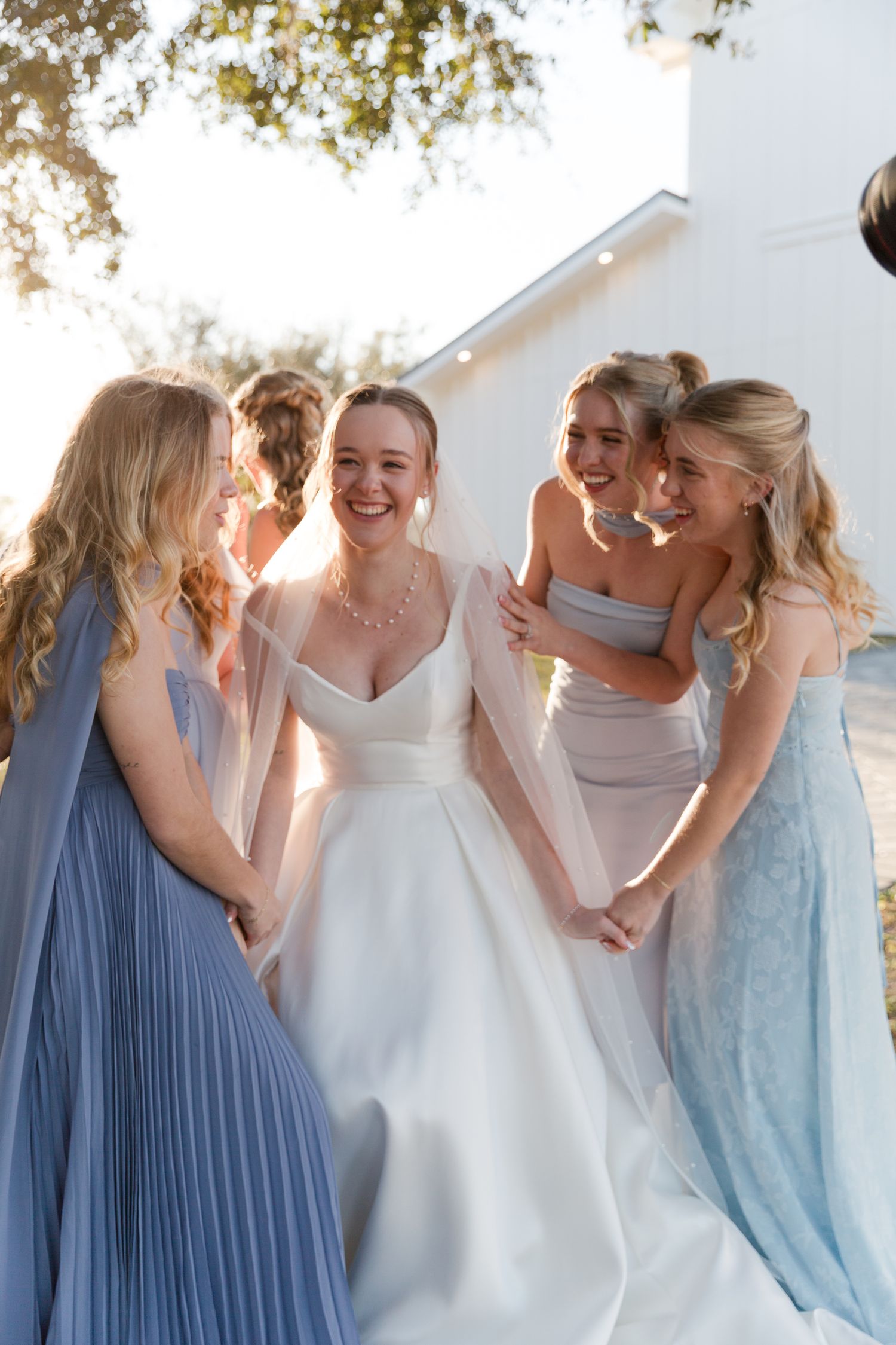 Bride in white gown laughing with four bridesmaids in blue and gray dresses outdoors on a sunny day.