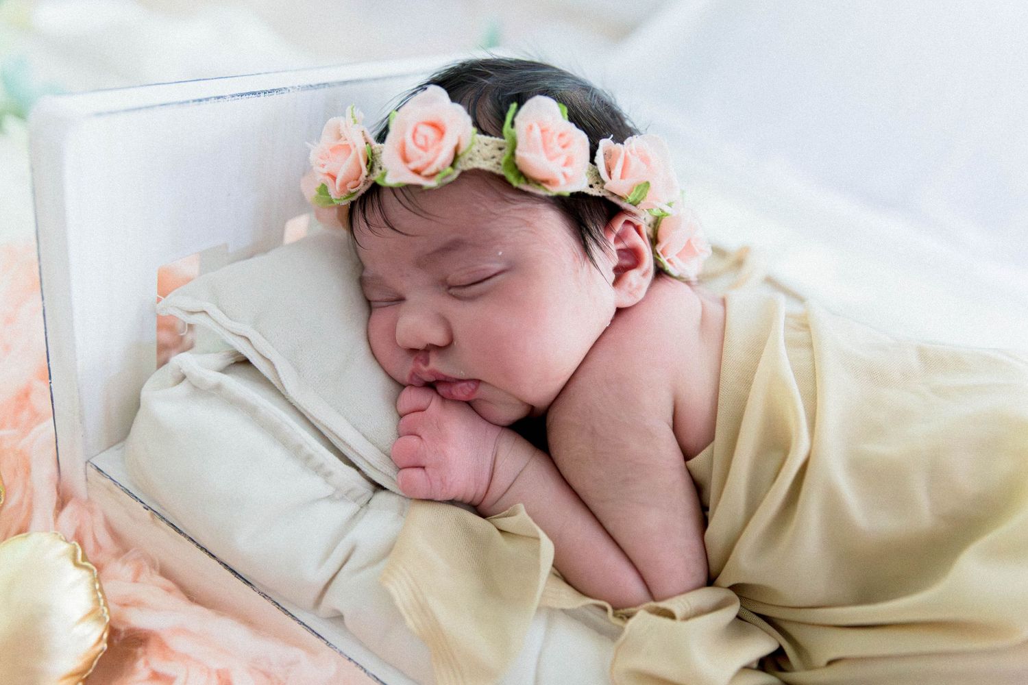 Newborn baby sleeping peacefully wearing a pink floral crown, wrapped in a soft golden blanket in a white wooden box.