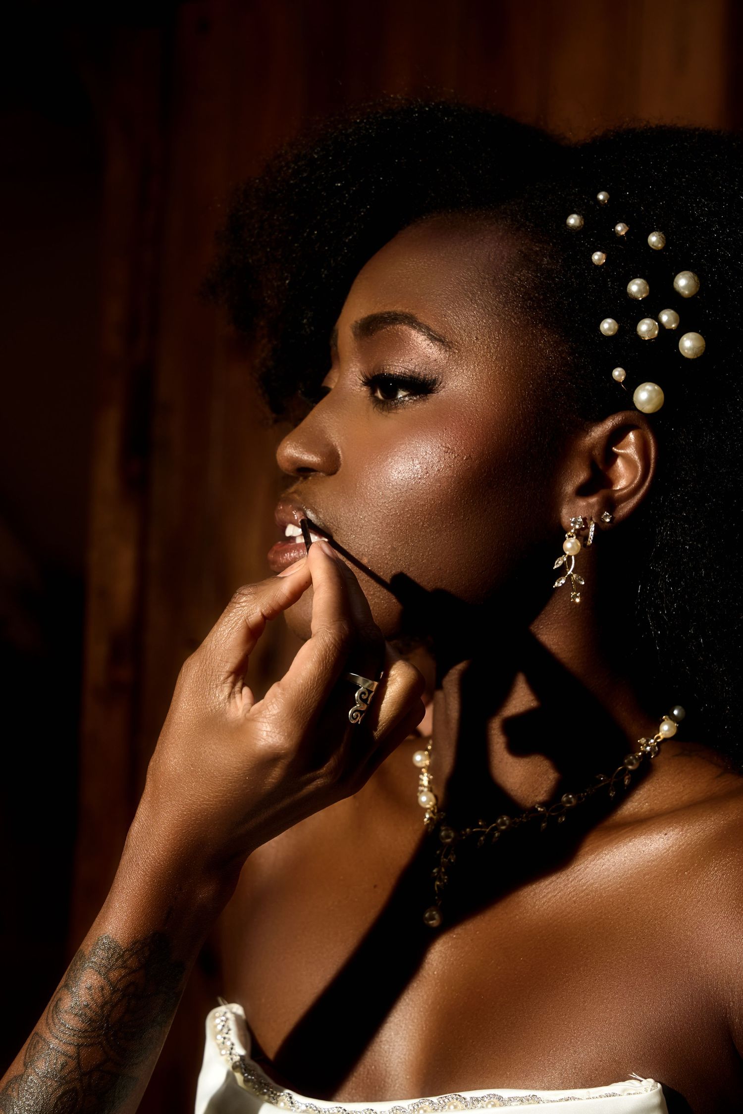 Black woman with pearl hair accessories applying lip makeup, wearing gold jewelry and a black ribbon choker.