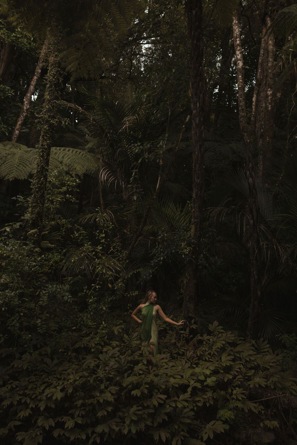 Woman in green dress standing among lush tropical rainforest trees and dense green foliage.