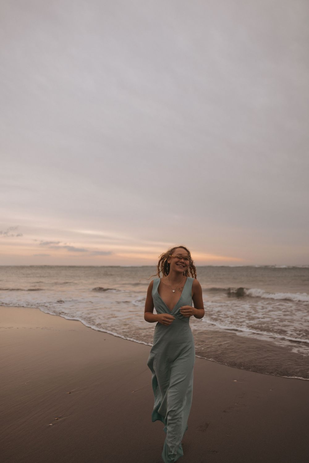 A smiling woman in a light green dress walks along a sandy beach at sunset with ocean waves behind her.