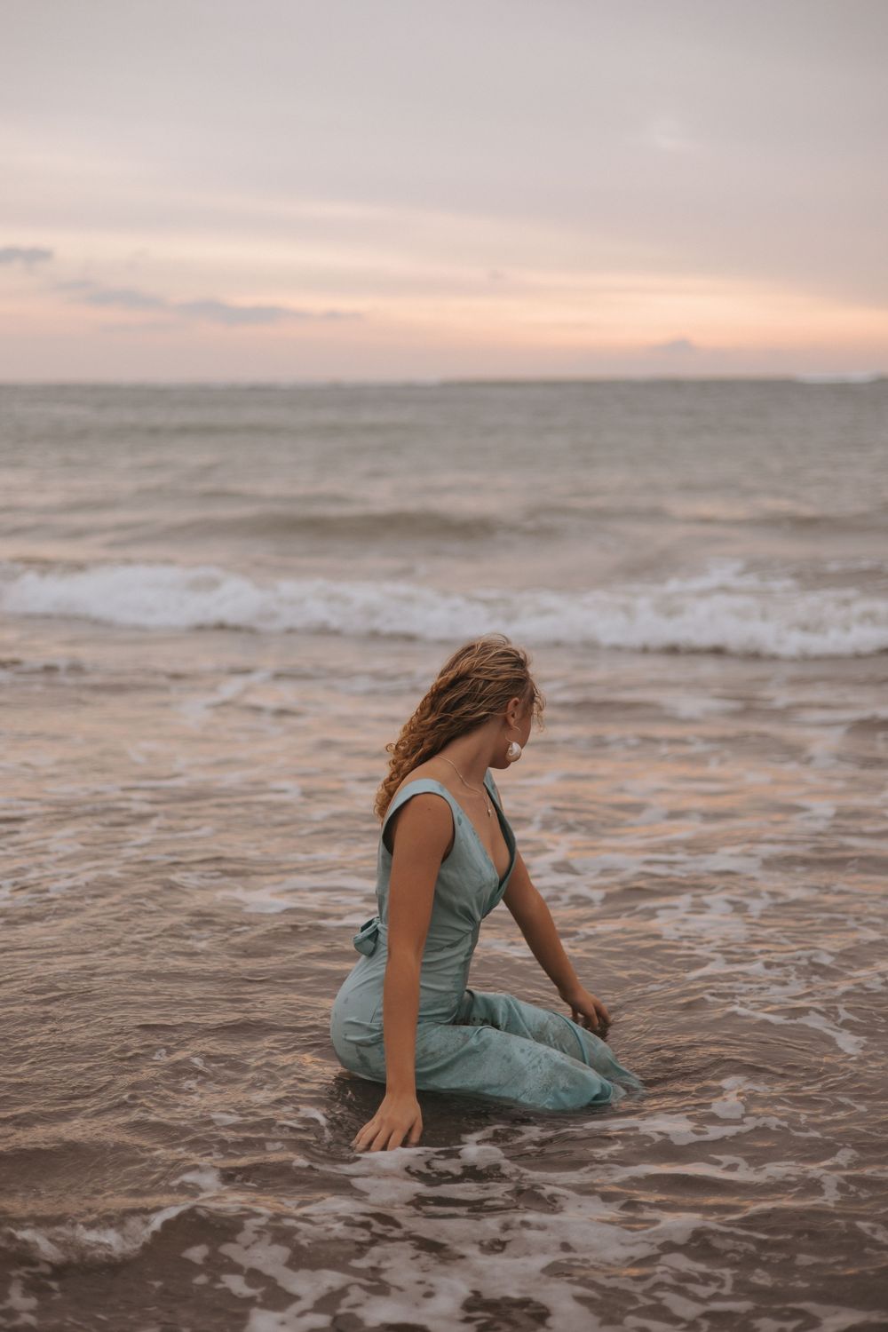 Woman in teal jumpsuit sitting on sandy beach shore with ocean waves and pastel sunset sky in background.