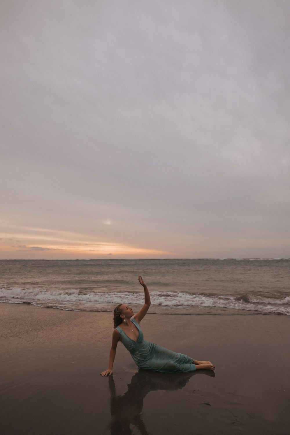 Woman in teal dress poses gracefully on a beach at sunset, with waves and dramatic cloudy sky in the background.