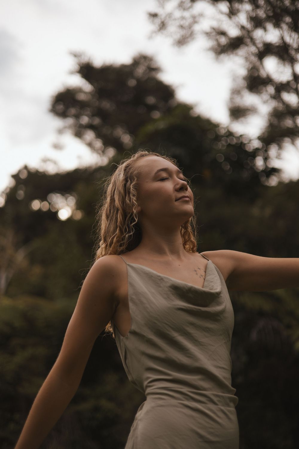 Young woman with wet curly hair wearing a beige slip dress, standing outdoors with eyes closed among lush green trees.