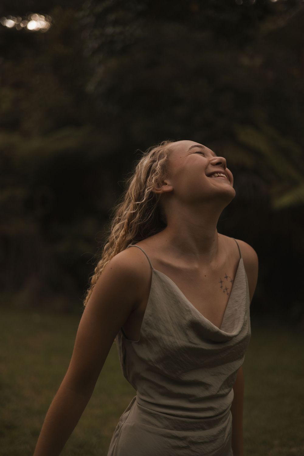 A joyful young woman with wavy blonde hair laughs outdoors, wearing a silver slip dress against a lush green background.