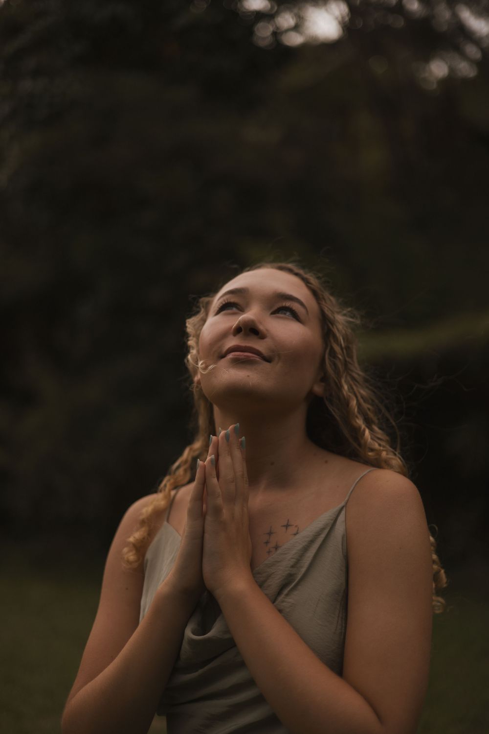 Young woman with curly hair praying outdoors, hands clasped together, looking up with a peaceful smile in a dark forest.