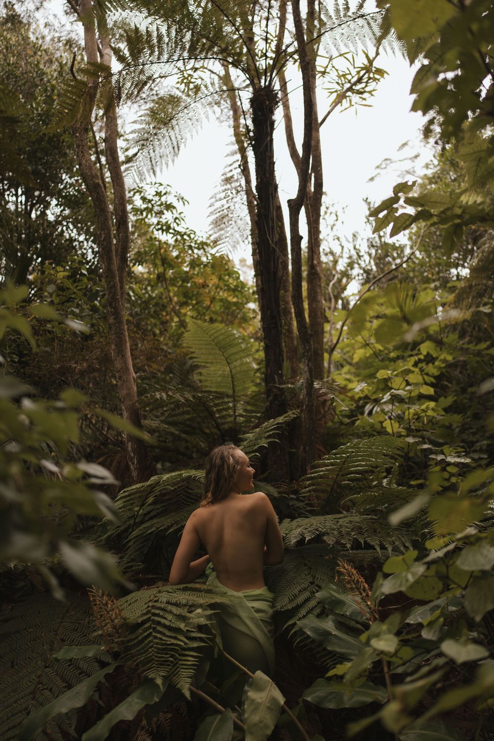 A shirtless person surrounded by lush green ferns and tropical trees in a dense jungle forest.