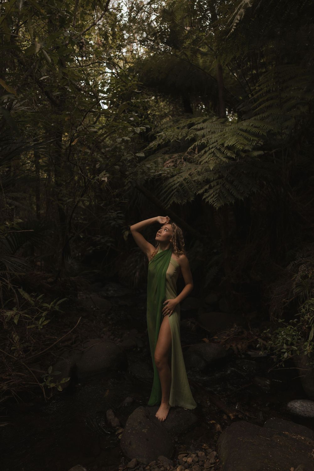 Woman in green draped dress standing on rock in lush dark forest with ferns.