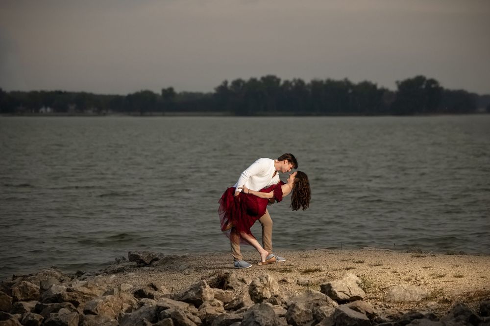 Couple in a romantic dance dip on a rocky lakeshore at dusk, the woman in a flowing red skirt, with calm water and distant trees behind.