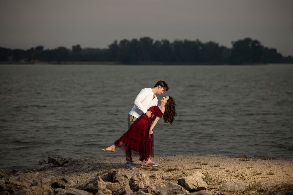 Couple shares a romantic dip kiss on a rocky lakeshore at sunset, the woman in a red dress and the man in a white shirt.