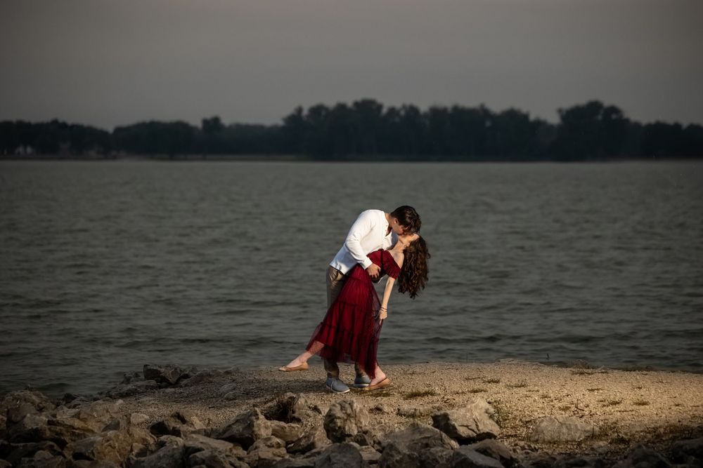 Couple in a romantic dip kiss on a rocky lakeshore at sunset, the woman in a flowing red dress, with calm water and trees behind.