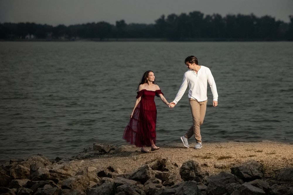 Couple holding hands and walking along a rocky lakeshore at sunset, she in a flowing burgundy dress and he in a white shirt and khaki pants.