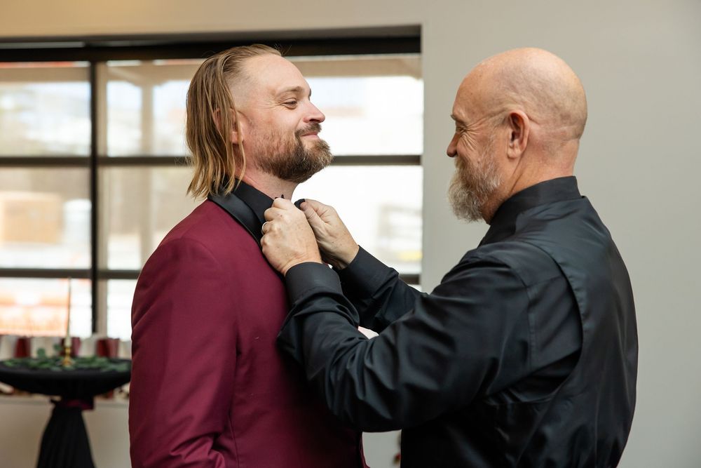 A bald, bearded man in a black shirt adjusts the bow tie of a smiling groom in a burgundy suit during wedding preparations.
